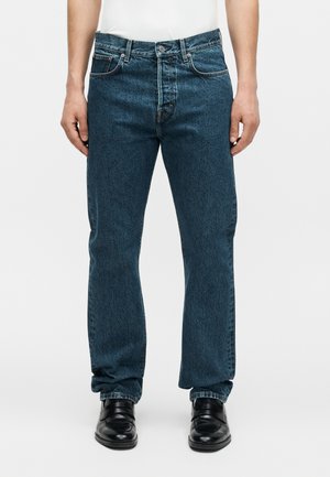 Άνδρας που φοράει σκουρόμπλε τζιν παντελόνια με ίσια πόδια, συνδυασμένα με μαύρα δερμάτινα παπούτσια και λευκό πουκάμισο που έχει μπει μέσα, στέκεται μπροστά σε απλό φόντο.