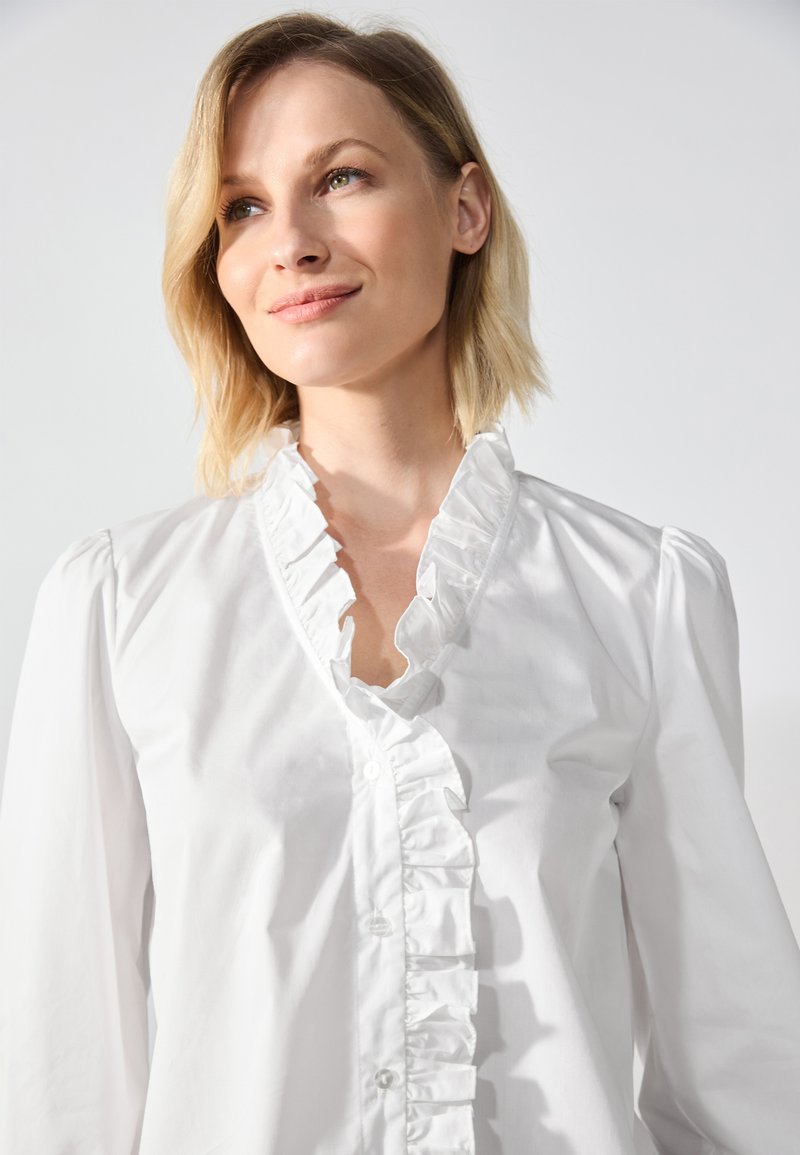 Blouse en coton blanc avec un col volanté, un boutonnière, et des manches longues avec des poignets froncés. Texture lisse, design classique.
