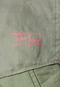 Tela verde claro con texto bordado en rosa "Hecho por Scotch & Soda AMS" y costuras visibles a lo largo de los bordes.