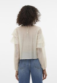 Blouse beige clair avec des manches à volants, en tissu transparent, et un dos en forme de trou de serrure. Associée à un jean en denim bleu taille haute.