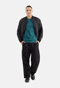 Chaqueta bomber negra con frontón texturizado, sudadera azul-verde con logotipo en relieve y pantalones cargo negros sueltos. Combinados con botas oscuras.