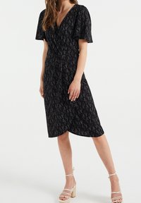 Kvinna som bär en svart omlottklänning med silverfärgad paisleymönster och volangärmar, tillsammans med vita sandaletter med blockklack och remmar.