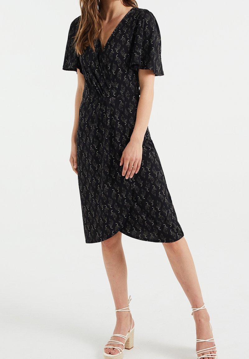 Kvinna som bär en svart omlottklänning med silverfärgad paisleymönster och volangärmar, tillsammans med vita sandaletter med blockklack och remmar.