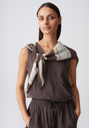 Woman with long dark hair wears sleeveless brown top and matching pants, accessorized with a large beige and brown striped scarf tied around her neck.