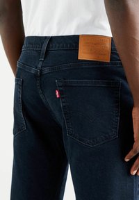 Mörkblå denimjeans med en läderplatta baktill, med klassiska sömmar och en liten röd flik på den vänstra bakfickan.