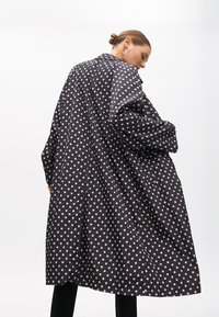 Mustapilkullinen takki, joka on valmistettu kevyestä kankaasta. Takissa on kaulus ja pitkät hihat sekä löysä istuvuus, joka laskeutuu lantioon asti.