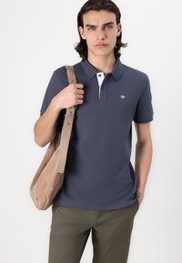 Marineblå polo shirt med hvid krave, korte ærmer og logo på brystet. Modellen bærer en lysebrun ruskinds skuldertaske.