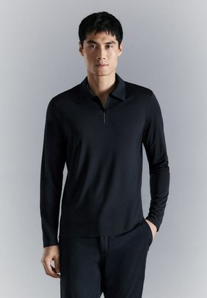 Joven de pie contra una pared lisa, vistiendo una camisa negra de manga larga con cuello de cremallera parcial y pantalones oscuros.