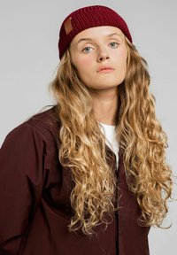 Burgundowa robiona na drutach czapka z prążkowanym wzorem i skórzaną naszywką z logo, noszona z ciemną, zapinaną na guziki kurtką na białą koszulę.