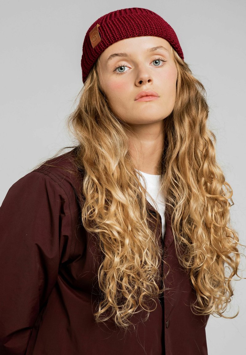 Burgundowa robiona na drutach czapka z prążkowanym wzorem i skórzaną naszywką z logo, noszona z ciemną, zapinaną na guziki kurtką na białą koszulę.