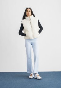 Bijela prsluk s visokim ovratnikom, prednjim patentnim zatvaračem i bočnim džepovima. Uparen s svijetloplavim hlačama širokih nogavica i bijelim debelim tenisicama.