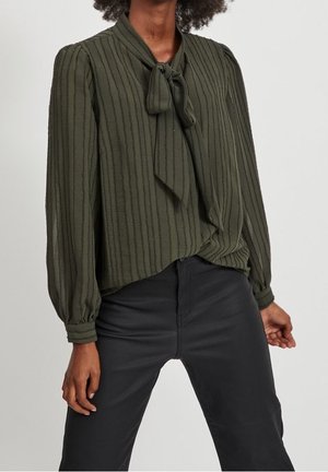 Vrouw draagt een olijfgroene blouse met verticale strepen en een gestrikte strik bij de hals, gecombineerd met zwart, hooggetailleerd broek.