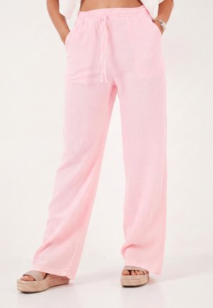 Persoon draagt een losse lichtroze broek met elastische tailleband en trekkoord, gecombineerd met beige platformsandalen, staat met handen in de zakken.