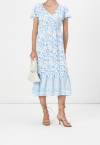 Robe florale paisley bleu clair avec un décolleté en V, des manches courtes volantées et un ourlet en niveaux. Associée à des sandales beige à lanières et un sac à main crème.