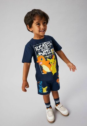 Smilende ung dreng iført navyblåt Pokémon-outfit med Charmander og Pikachu, stående mod en ensfarvet grå baggrund.