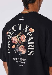 Camiseta de algodón negra con gran bordado floral en tonos de rosa y naranja, y texto en blanco que dice "PROJECT X PARIS" y "WILD SPIRIT PXP COLLECTION."