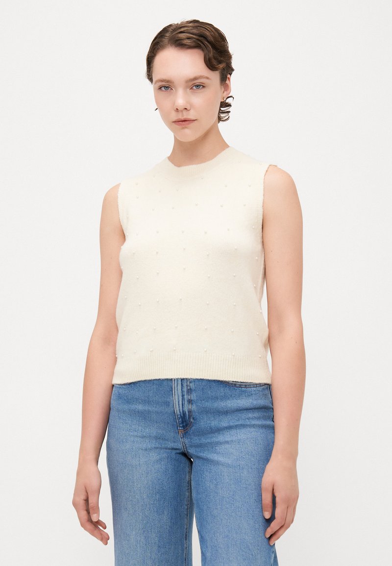 Uten ermer kremfarget strikket topp med rund hals og teksturert overflate med små, jevnt fordelte detaljer. Kombinert med blå denimjeans.