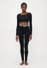 Frau in schwarzem, langärmeligen, kurzen Oberteil und schwarzen Leggings mit Moschino-Logo-Gummibund, barfuß vor einem schlichten Hintergrund stehend.