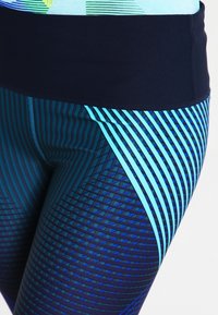 Leggings bleus à motifs avec une taille sombre, présentant des lignes diagonales et verticales dans des nuances variées de bleu. Tissu lisse.