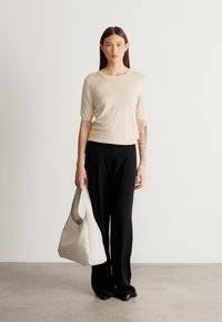 Bluză bej cu mâneci scurte, pantaloni negri cu picioare largi, ținând o geantă mare de umăr crem. Modelul stă pe un fundal neutru, cu podea netedă.