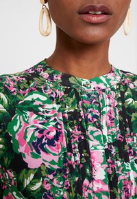 Blusa floral con patrones en verde, rosa y negro, textura plisada y un cuello redondo con detalle de botón. Pendientes circulares grandes.