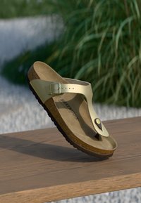Goldene Zehensandalen mit einem glatten, synthetischen Obermaterial, verstellbarem Schnallenverschluss, Korkfußbett und rutschfester Gummisohle. Markenname auf dem Fußbett sichtbar.
