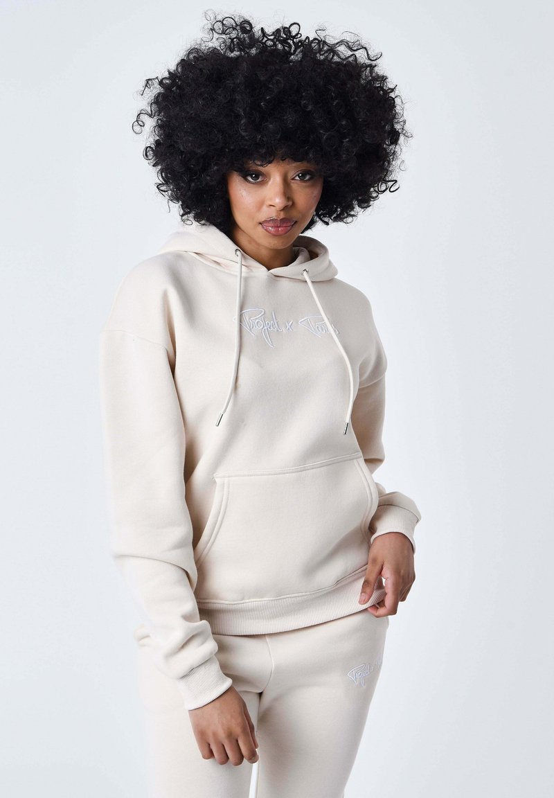 Hoodie beige realizzato in tessuto morbido con tasca frontale, cordini e logo ricamato. Abbinato a pantaloni coordinati, con un design simile.