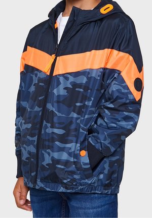 Kind trägt eine marineblaue und orange Tarnjacke mit Frontreißverschluss und Kapuze, Hände in den Taschen, kombiniert mit blauen Jeans.