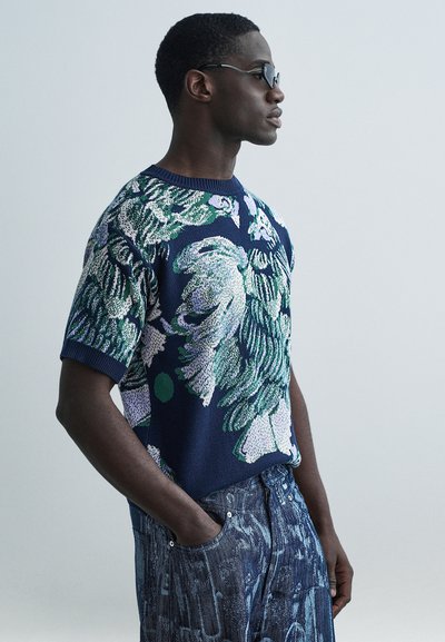 Pull en maille bleu marine avec motif floral vert et blanc, manches courtes et col rond côtelé. Associé à un pantalon en denim foncé avec poches.