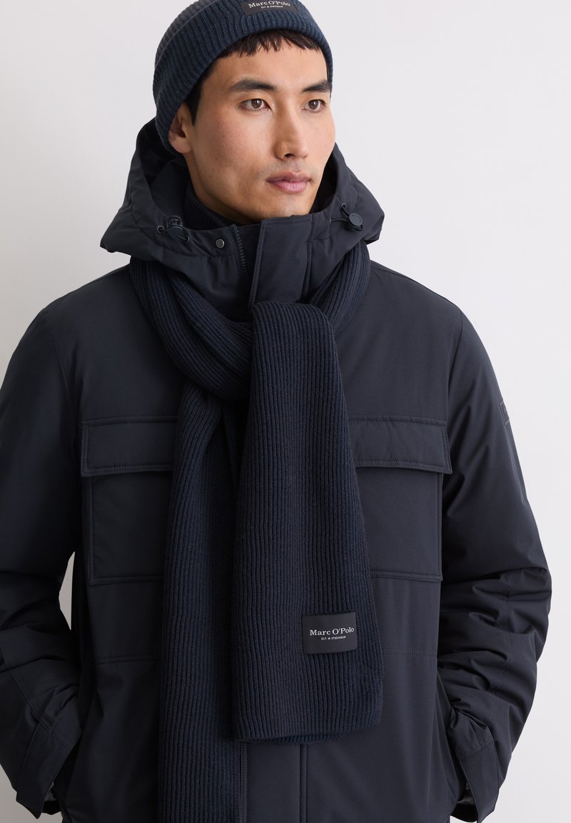 Marine blauwe gebreide sjaal met ribbeltextuur, voorzien van een zwart label. Draagt bij een bijpassende beanie en een donkere jas, wat het ontwerp van winterkleding benadrukt.