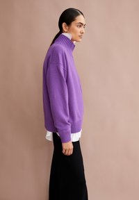 Frau, die im Profil steht und einen lila Strickpullover über einem weißen Hemd und schwarzen Hosen trägt, vor einem schlichten beige Hintergrund.
