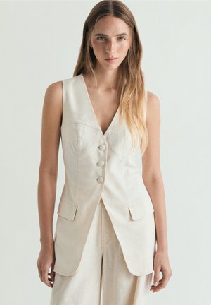 Mujer con cabello castaño claro y pecas que lleva un chaleco crema sin mangas con botones y pantalones de pierna ancha a juego sobre un fondo liso.