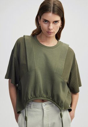 T-shirt cropped vert olive avec des panneaux texturés, col rond et cordon de serrage réglable à l'ourlet. Tissu doux avec une coupe décontractée.