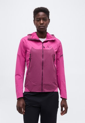 Femme portant une veste zippée bicolore rose avec capuche et un pantalon noir, debout devant un fond clair uni.