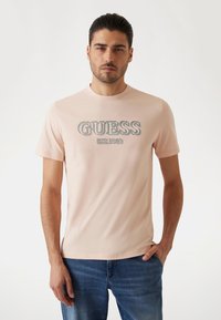 Camiseta de manga corta de color rosa claro con "GUESS EST. 1981" en letras grises. Corte regular, textura suave, combinada con jeans de mezclilla azul.