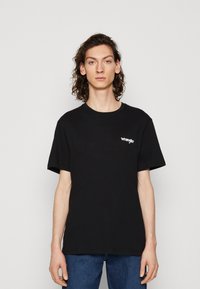 Črna bombažna majica s sproščenim krojem in okroglim izrezom. Na prsih ima bel logo »Wrangler«.