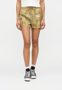 Femme portant un short imprimé tropical vert olive, des chaussettes blanches et des baskets montantes noires, debout contre un fond blanc.