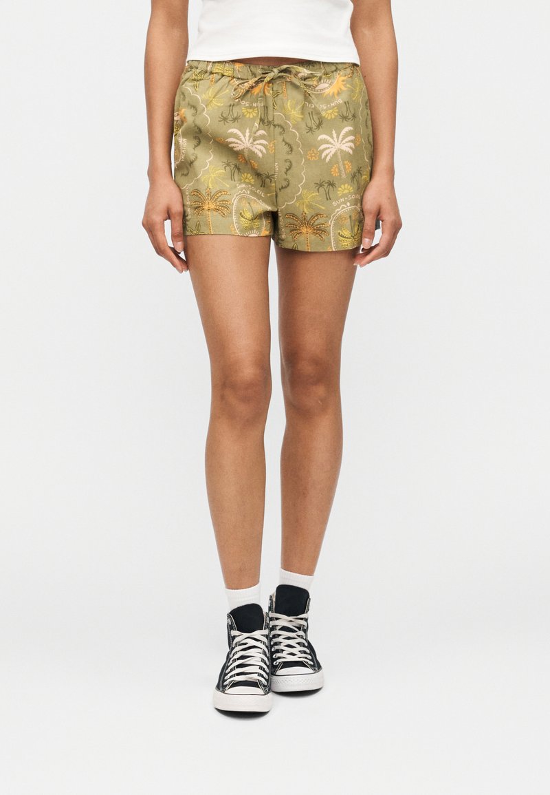 Femme portant un short imprimé tropical vert olive, des chaussettes blanches et des baskets montantes noires, debout contre un fond blanc.