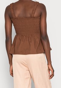 Haut péplum smocké marron avec bretelles volantées, présentant un corsage texturé et un ourlet évasé. Associé à un pantalon ample beige clair.