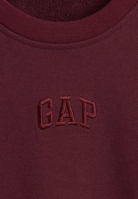 Bordo dukserica s okruglim izrezom i mekanom teksturom, s izvezenim logotipom "GAP" u sredini. Jednostavan i neukrašen dizajn.