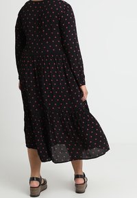 Robe noire à manches longues, design en couches, motif floral rouge et ourlet fluide. Portée avec des sandales à plateforme noires.