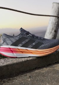 Chaussure de course grise et noire avec une semelle intermédiaire dégradée orange et rose reposant sur un rebord en béton au coucher du soleil à l'extérieur.