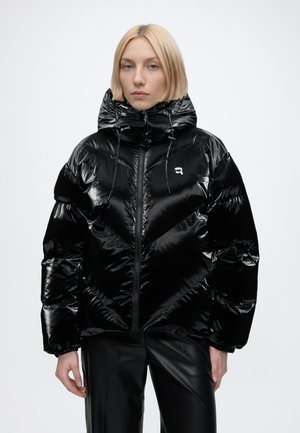 Femme blonde portant une veste matelassée noire brillante avec une capuche et un pantalon noir, debout devant un fond clair uni.