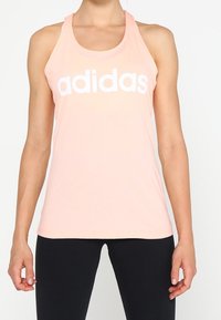 Top deportivo de color rosa claro, confeccionado con una tela suave. Presenta un gran logo blanco de "adidas" en el frente y un escote en forma de barco.