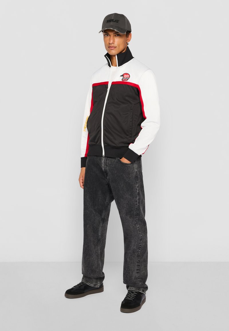 Schwarze und weiße Zip-Jacke mit roten Akzenten, elastischen Bündchen und besticktem Logo, kombiniert mit dunkelblauen Jeans und schwarzen Turnschuhen.