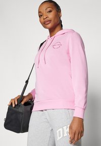 Helles rosa Hoodie mit einer Vordertasche, Kapuze mit Kordelzug, gerippten Bündchen und Saum, kombiniert mit grauen Jogginghosen. Schwarze Tasche mit Schulterriemen wird in der Hand gehalten.