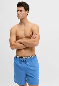 Homme torse nu aux cheveux courts et foncés, debout les bras croisés, portant un short de bain bleu avec une ceinture noire étiquetée "JACK & JONES."