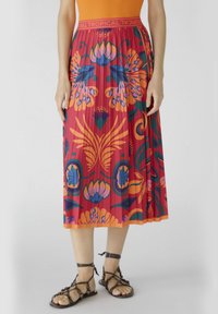 Gonna plissettata rossa con motivi floreali tropicali in arancione, blu e verde. Vita elastica con testo "Tropical". Abbinata a sandali con cinturini.