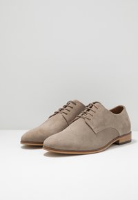 Chaussures oxford en suédine beige avec un bout pointu, cinq œillets et un petit talon en bois brun. Texture lisse avec des accents de design minimalistes.