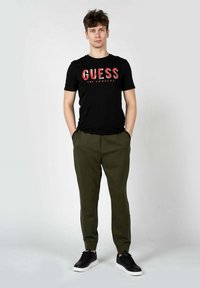 Sort bomulds T-shirt med rød "GUESS LOS ANGELES" grafik, parret med olivengrønne tapered bukser og sorte sneakers.